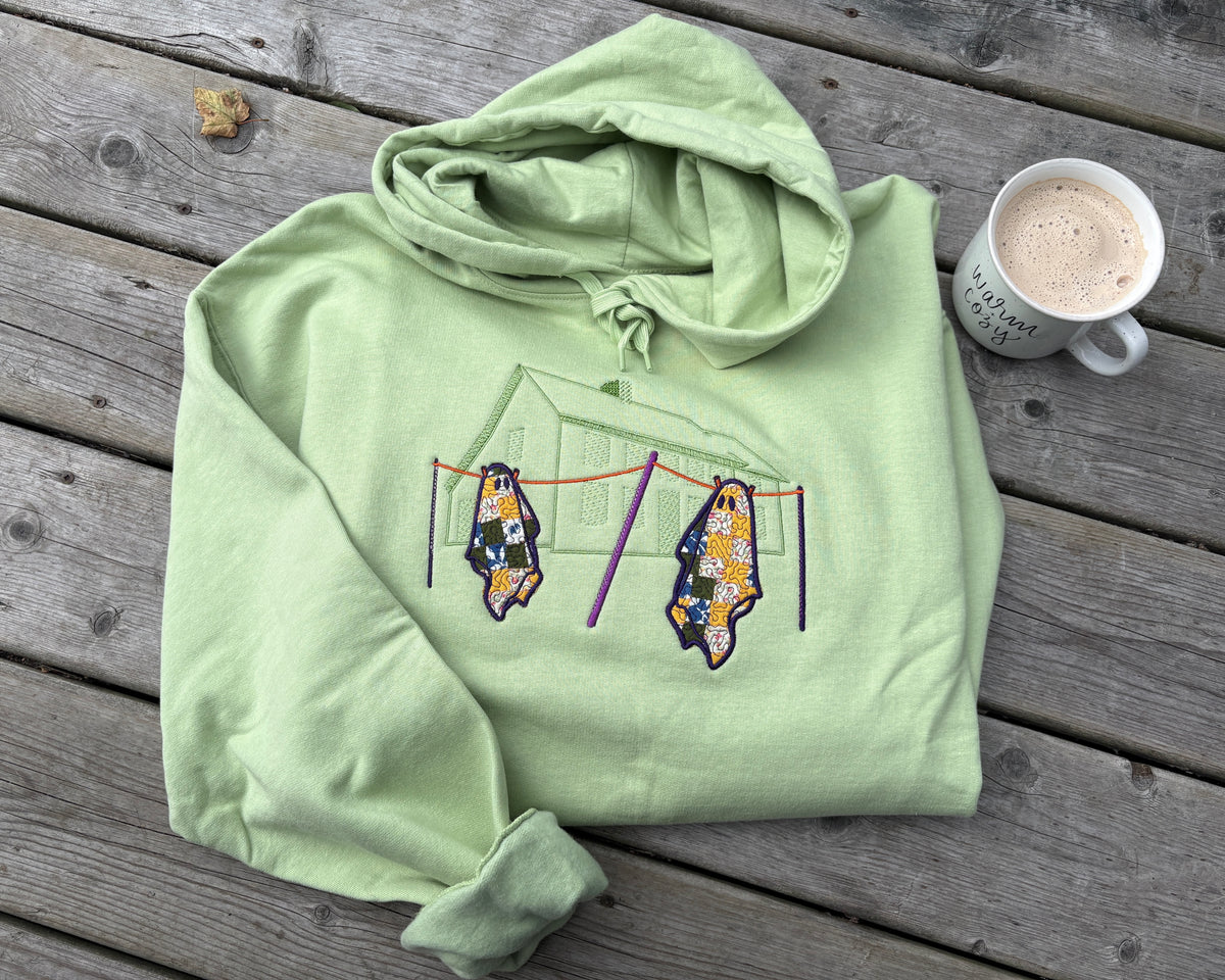 A pistachio green folded hoodie resting on a wooden deck. The design on the front of the hoodie is a outline of a saltbox house stitched tone on tone in green stitching. In the foreground of the stitched design there is a clothes line with the poles stitched in dark purple and the line stitched in orange. Hanging on the line are two quilted ghosts with the quilts in various colours with a purple swirl stitched overtop. To the right of the hoodie there is a white mug holding a latte that says "warm & cozy".