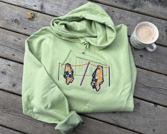 A pistachio green folded hoodie resting on a wooden deck. The design on the front of the hoodie is a outline of a saltbox house stitched tone on tone in green stitching. In the foreground of the stitched design there is a clothes line with the poles stitched in dark purple and the line stitched in orange. Hanging on the line are two quilted ghosts with the quilts in various colours with a purple swirl stitched overtop. To the right of the hoodie there is a white mug holding a latte that says "warm & cozy".