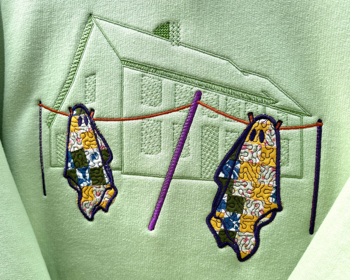 Pistachio green fabric showing a close up of the design on the front of the hoodie. There is an outline of a saltbox house stitched tone on tone in green stitching. In the foreground of the stitched design there is a clothes line with the poles stitched in dark purple and the line stitched in orange. Hanging on the line are two quilted ghosts with the quilts in various colours with a purple swirl stitched overtop.