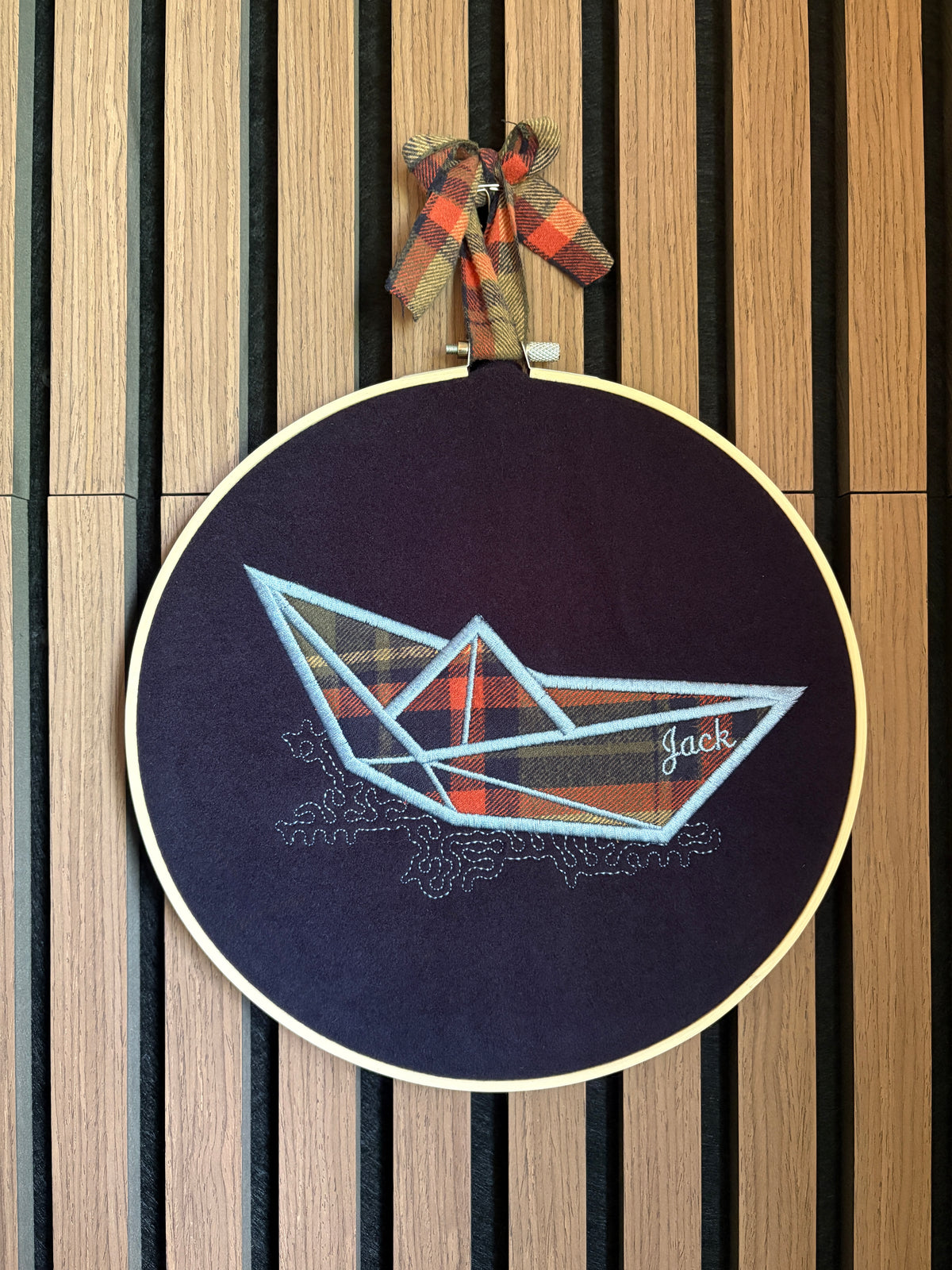A wooden circular embroidery hoop hanging on a slatted wood wall, featuring an origami paper boat design. The boat is filled with a plaid fabric with the outline stitched in light blue. Beneath the boat are squiggles representing the water and the name 'Jack' is embroidered on the hull of the boat. The same plaid shirt fabric is used to make a tie from which it hangs.